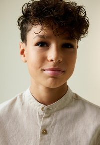 Fiatal személy portréja, rövid, göndör barna hajjal és semleges arckifejezéssel, világos bézs mandzsettás ingben, mandarin gallérral.