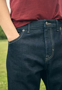 Persoon draagt donkerblauwe spijkerbroek met hand in voorzak en een rode t-shirt losjes in de tailleband gestopt.