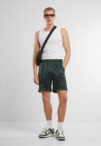 Shorts de sport verts avec ceinture élastique, débardeur blanc, sac bandoulière noir et baskets vertes avec accents blancs. Lunettes de soleil sombres portées.