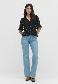 Schwarze Bluse mit floralem Muster und Rüschenärmeln, verziert mit blauen Blumenmustern, kombiniert mit hellblauen Schlagjeans und braunen Stiefeletten mit Absatz.