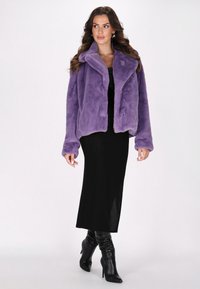 Giacca in pelliccia sintetica viola con colletto, apertura frontale e maniche lunghe, abbinata a un vestito nero e stivali neri fino al ginocchio.