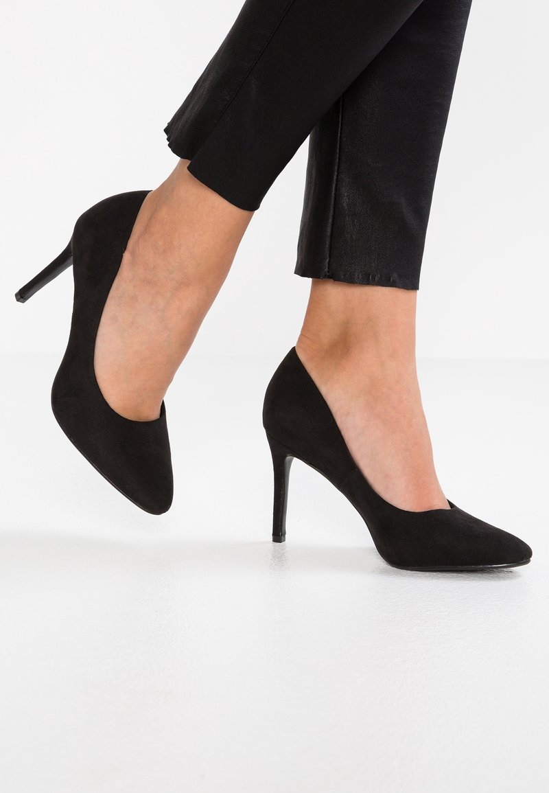 Frau trägt schwarze Wildleder-Pumps mit spitzem Zehenbereich und schmalen Stilettos, kombiniert mit schwarzen Cropped-Hosen vor einem weißen Hintergrund.