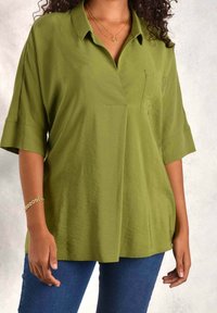 Olive green short-sleeve shirt with a collar and front pocket, featuring a loose fit and smooth texture. Worn with blue jeans.