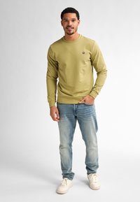Sudadera verde oliva de manga larga y cuello redondo, con un pequeño logo en el pecho. Combinada con jeans de tono azul claro y zapatillas blancas.
