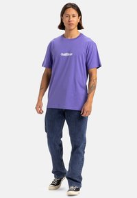 Camiseta de algodón morada de mangas cortas con logo blanco en el pecho, combinada con pantalones vaqueros azules y zapatillas negras.