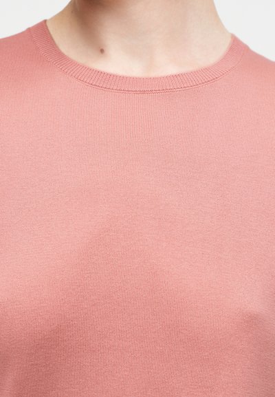 Κοντινό πλάνο ενός ατόμου που φοράει ένα απλό, απαλό ροζ πλεκτό πουλόβερ με στρογγυλή λαιμόκοψη, δείχνοντας τον λαιμό και το ανώτερο μέρος του στήθους.