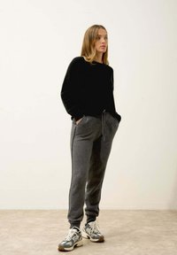 Černý pletený svetr, šedé fleecové tepláky s bočními kapsami a šňůrkou, doplněné sportovními teniskami v šedých a bílých tónech.