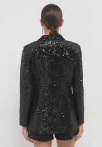 Donna con capelli raccolti, che indossa un blazer nero con paillettes e pantaloni corti abbinati, in piedi di spalle su uno sfondo uniforme.