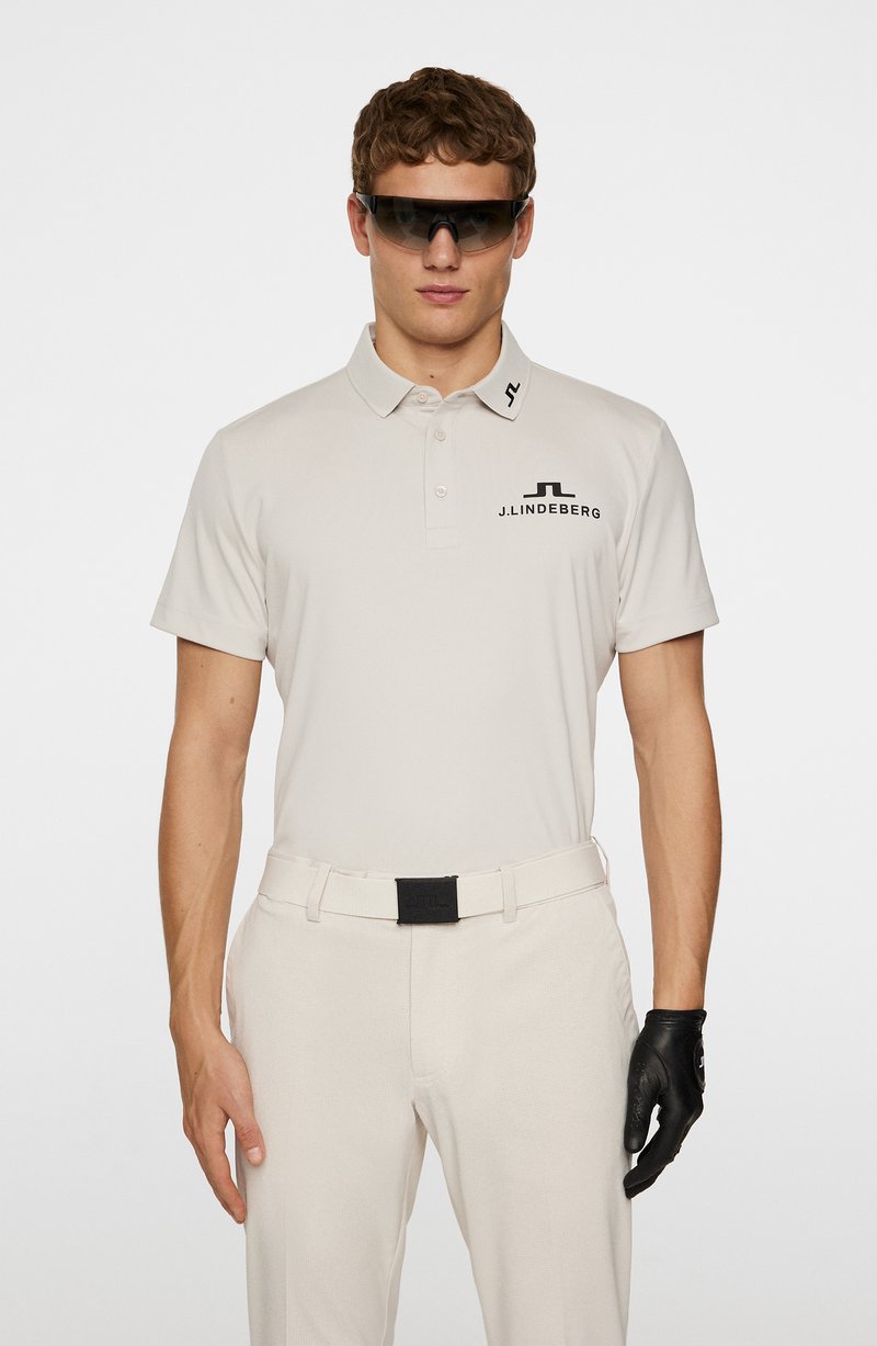 Polo-shirt beige clair à manches courtes et col, avec un logo noir. Associé à un pantalon beige clair et une ceinture noire. Gant noir porté à la main droite.