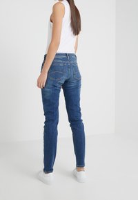 Tmavě modré skinny džíny s zadními kapsami, lehkým opotřebením a středním pasem, v kombinaci s bílým bezrukávovým topem a bílými teniskami.