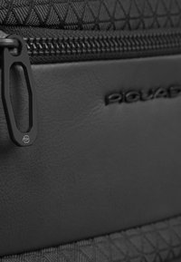 Tessuto nero texturizzato con un motivo geometrico, dettagli in pelle liscia e una cerniera metallica con dettaglio logo.