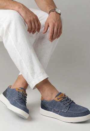 Mann trägt weiße Hose und blaue lässige Schnürschuhe mit weißen Sohlen, sitzt mit gekreuzten Beinen und die Hände auf den Knien ruhend.