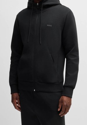 Schwarzer Kapuzenpullover mit Reißverschluss, Kordeln und Seitentaschen, kleines "BOSS"-Logo auf der linken Brust, getragen von einer Person vor einfachem Hintergrund.