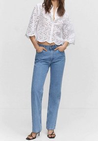 Crop top blanc en broderie anglaise avec ourlet en forme de coquille, associé à un jean bleu taille haute. Des sandales noires à brides complètent la tenue.