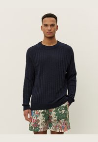 Granatowy sweter z prążkowanej dzianiny z raglanowymi rękawami i okrągłym dekoltem. W połączeniu z wzorzystymi szortami z tropikalnymi i ludzkimi ilustracjami.