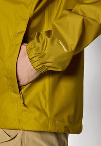 La veste imperméable jaune présente un poignet élastique et une poche latérale, mettant en valeur la marque DRYVENT. Texture de tissu lisse et légère.