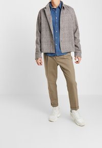 Grijze geruite jas over een blauwe blouse, beige broeken en witte sneakers. Opvallende textuur in de stof van de jas.