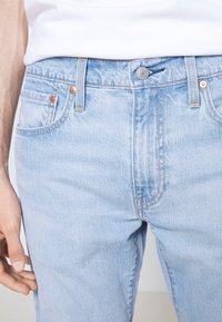 Gros plan d'une personne portant un jean en denim bleu clair et une chemise blanche, montrant la taille, le bouton et les poches avant.