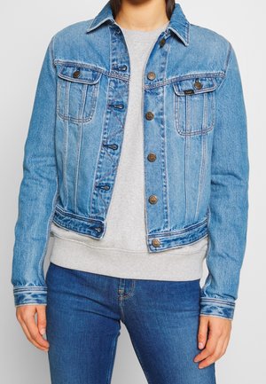 Person iført blå Lee denimjakke over lysegrå sweatshirt og blå jeans, stående mod en ensfarvet grå baggrund.