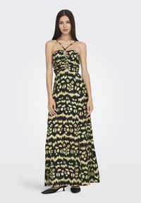 Mujer con un vestido largo sin mangas de color negro y amarillo con un estampado, de tirantes entrelazados, de pie contra un fondo claro y liso.