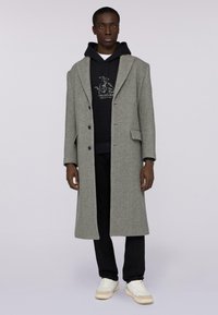 Szary wełniany płaszcz z klapami, na dwa guziki, z bocznymi kieszeniami, noszony na czarnym hoodie i czarnych spodniach, zestawiony z białymi trampkami.