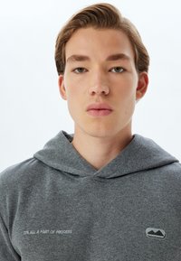 Sudadera gris hecha de tela suave con capucha, que presenta un logo y el texto "TODO FORMA PARTE DEL PROCESO" bordado en blanco.
