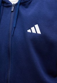 Σκούφο με φερμουάρ σε ναυτικό μπλε χρώμα με λευκό λογότυπο τριών γραμμών στο στήθος, με κορδόνι κοντά στο φερμουάρ.