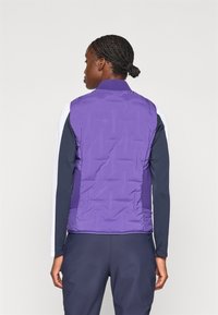 Gilet matelassé violet avec col côtelé, porté sur une chemise à manches longues bleu marine, présentant des panneaux latéraux et une texture lisse. Vue arrière présentée.