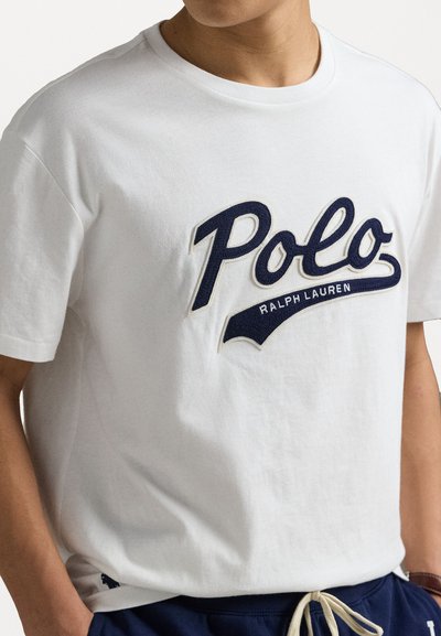 Balta kokvilnas T-kreklu, kas aprīkots ar tumši zilu izšūtu "Polo" logo un zem tā "Ralph Lauren", īsām piedurknēm un apaļu dekoltē.