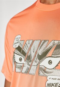 Kortærmet orange T-shirt med et grafisk design med sorte øjne og tekst, udført i en tegneseriestil, glat tekstur.