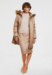 Beżowa kurtka puchowa z kapturem z sztucznego futra, połączona z jasnoróżowym swetrem w warkocze i beżową spódnicą ołówkową. Całość dopełniają neutralne obcasy.