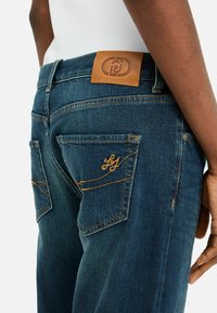 Jeans de denim en azul oscuro con una textura suave. Presenta dos bolsillos traseros, costuras en un tono amarillo claro y un parche de cuero marrón de la marca.