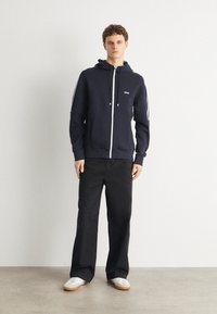Marinblå zip-up hoodie med dragsko på huvan och vita sidostråk. Har en liten logotyp på bröstet. Kombineras med svarta vidbyxor och sneakers.
