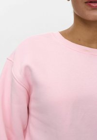 Gaiši rozā džemperis arapaļo izgriezumu. Raksturīga mīksta tekstūra un brīva piegriezuma forma. Redzami šuvju detaļas plecos.