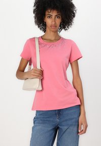 Pink short-sleeve t-shirt with a round neckline and decorative rhinestones along the shoulder. Paired with blue jeans, holding a beige bag.