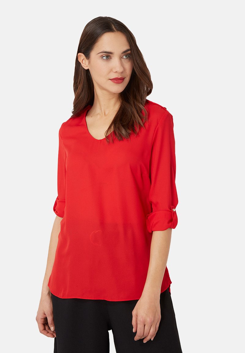 Mujer con cabello largo y castaño que lleva una blusa roja de cuello en V con las mangas arremangadas y pantalones negros, mirando ligeramente hacia la derecha sobre un fondo blanco.