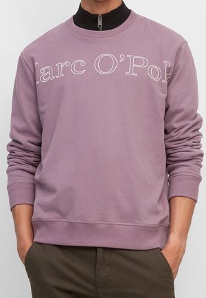 Osoba w mauve'owej (jasnofioletowej) bluzie z napisem „"Marc O'Polo"” na piersi, nałożonej na czarny sweter z zamkiem przy kołnierzu, ręce w kieszeniach brązowych spodni.
