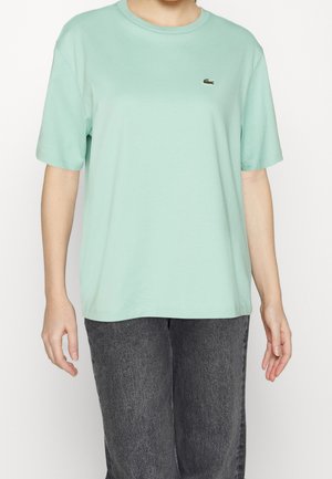 Osoba w miętowo zielonym t-shircie z krótkim rękawem, z małym ciemnozielonym logo krokodyla, oraz wyblakłych czarnych dżinsach, stojąca na białym tle.