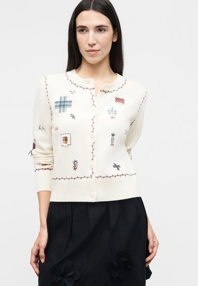 Γυναίκα που φοράει λευκή ζακέτα με κουμπιά και κεντημένες επιδιορθώσεις με κόκκινα ράμματα, σε συνδυασμό με μαύρη φούστα με υφασμάτινα λουλούδια.