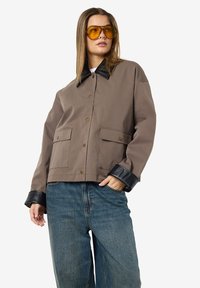 Vrouw in een grijze jas met een zwarte leren kraag en manchetknopen, met geel-getinte zonnebril en blauwe jeans, staat met één hand in haar zak.