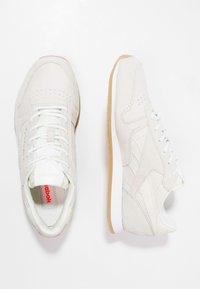 Ett par vita Reebok-sneakers med mockatextur, vita snören och ljusbruna gummisålar på en vit bakgrund.