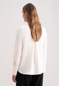 Cream-colored blouse with long sleeves, soft fabric, and buttoned cuffs. Back features a single pleat and a smooth, tailored design.
