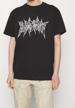Person iført en sort T-shirt med hvid takkede tekstgrafik og lyse beige cargoshorts mod en ensfarvet hvid baggrund.