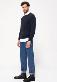 Granatowy sweter z prążkowanej dzianiny, biała koszula z kołnierzykiem noszona pod spodem, niebieskie drewniane dżinsy, oraz czarne sznurowane botki.
