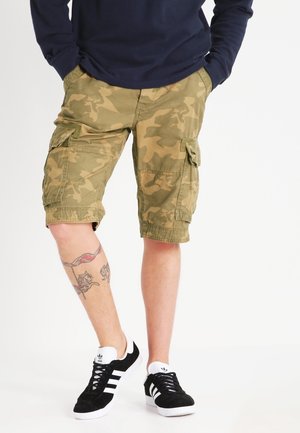 Pantalones cortos cargo de camuflaje en verde oliva con múltiples bolsillos, un ajuste relajado y detalles de costura, combinados con zapatillas negras y blancas.
