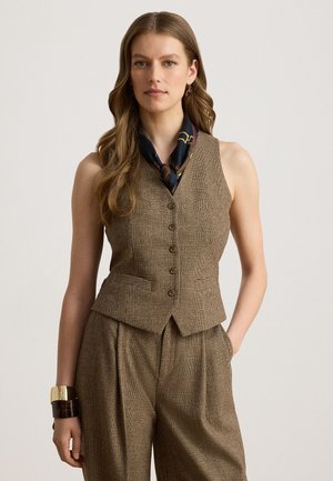 Frau trägt braune karierte Weste und passende hoch taillierte Hose mit einem dunkel gemusterten Halstuch und großen goldenen sowie Schildpatt-Armbändern.
