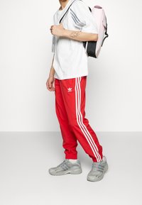 Röda träningsbyxor med vita sidostreck, en vit t-shirt med gråa ränder och grå sneakers. En rosa och svart ryggsäck hänger på axeln.