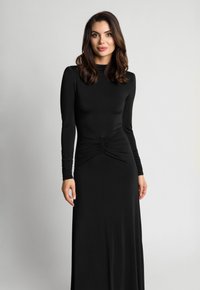 Kobieta z długimi brązowymi włosami, ubrana w dopasowaną czarną sukienkę z długim rękawem i marszczeniami w talii, stojąca na tle jednolitego, jasnego tła.