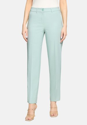 Frau trägt hellblaue, gerade geschnittene Hose mit Vorderfalte, Knopfverschluss und beigen offenen High Heels, steht vor neutralem Hintergrund.