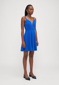 Blue sleeveless dress with a deep V-neck, fitted bodice, and flared skirt. Worn with black heeled shoes featuring decorative buckles.